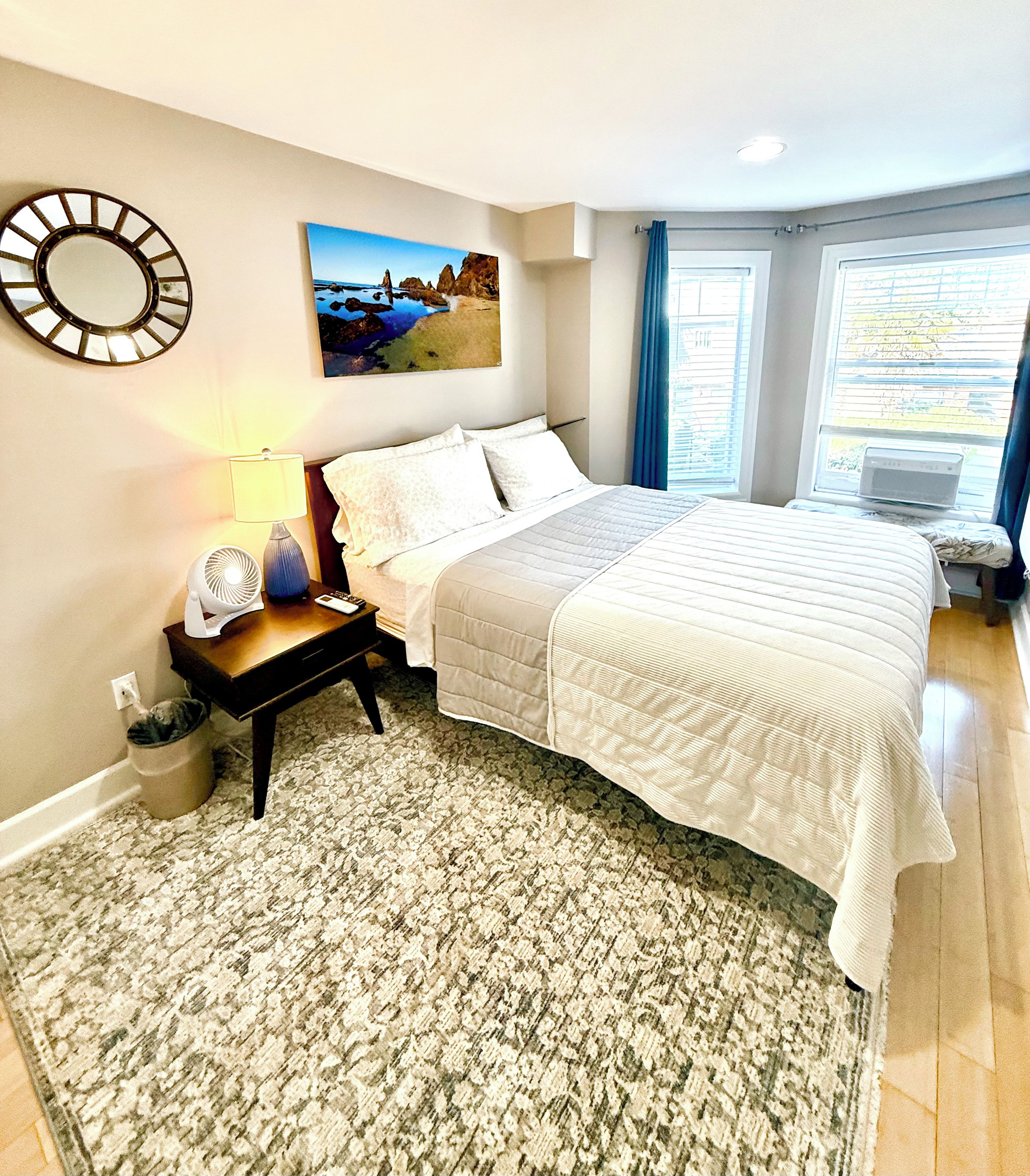 A bedroom with a bed, a nightstand, a lamp, a picture on the wall, and a window with curtains.