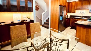 A modern kitchen with dark wooden cabinets, a stainless steel refrigerator, and a glass dining table with wicker chairs. - 180° Space Needle, Lake Union & Downtown Views! (Seattle)