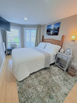 A bedroom with a large bed, a bedside table with a lamp, a wall-mounted TV, and a window with blinds. - 180° Space Needle, Lake Union & Downtown Views! (Seattle)