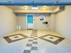 Espaço interior limpo e bem iluminado, com um padrão de piso xadrez, uma porta azul, uma lixeira para reciclagem e uma estação de carregamento na parede.