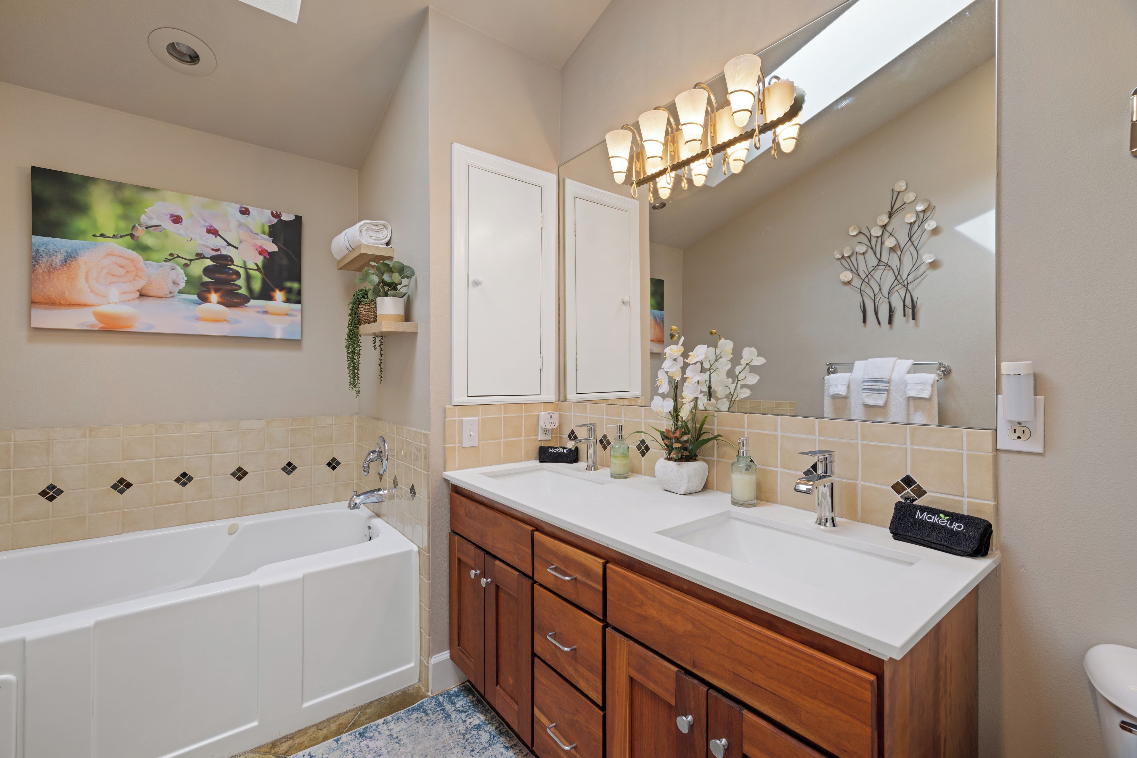 A bathroom with a bathtub, two sinks, wooden cabinets, a large mirror, and a wall-mounted light fixture.