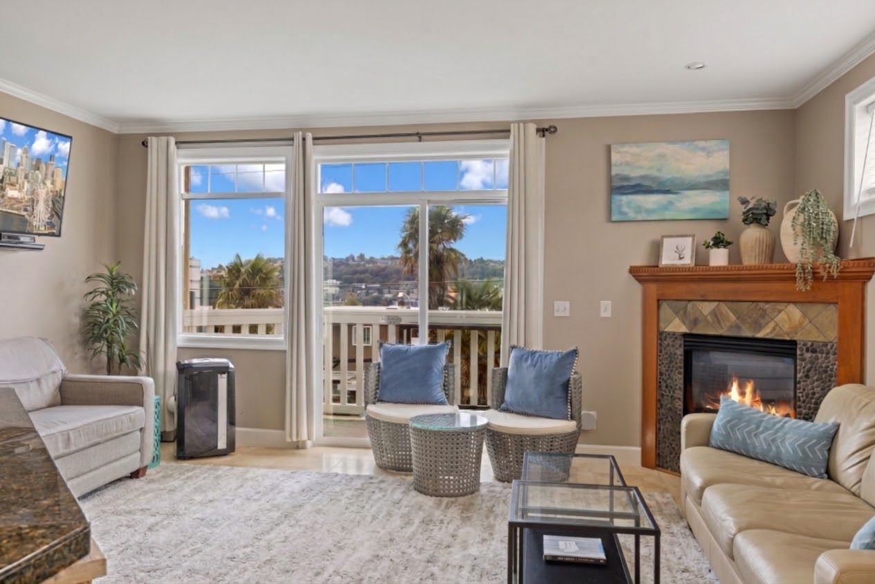 A living room with a fireplace, a sofa, a coffee table, and a view of a cityscape through large windows.