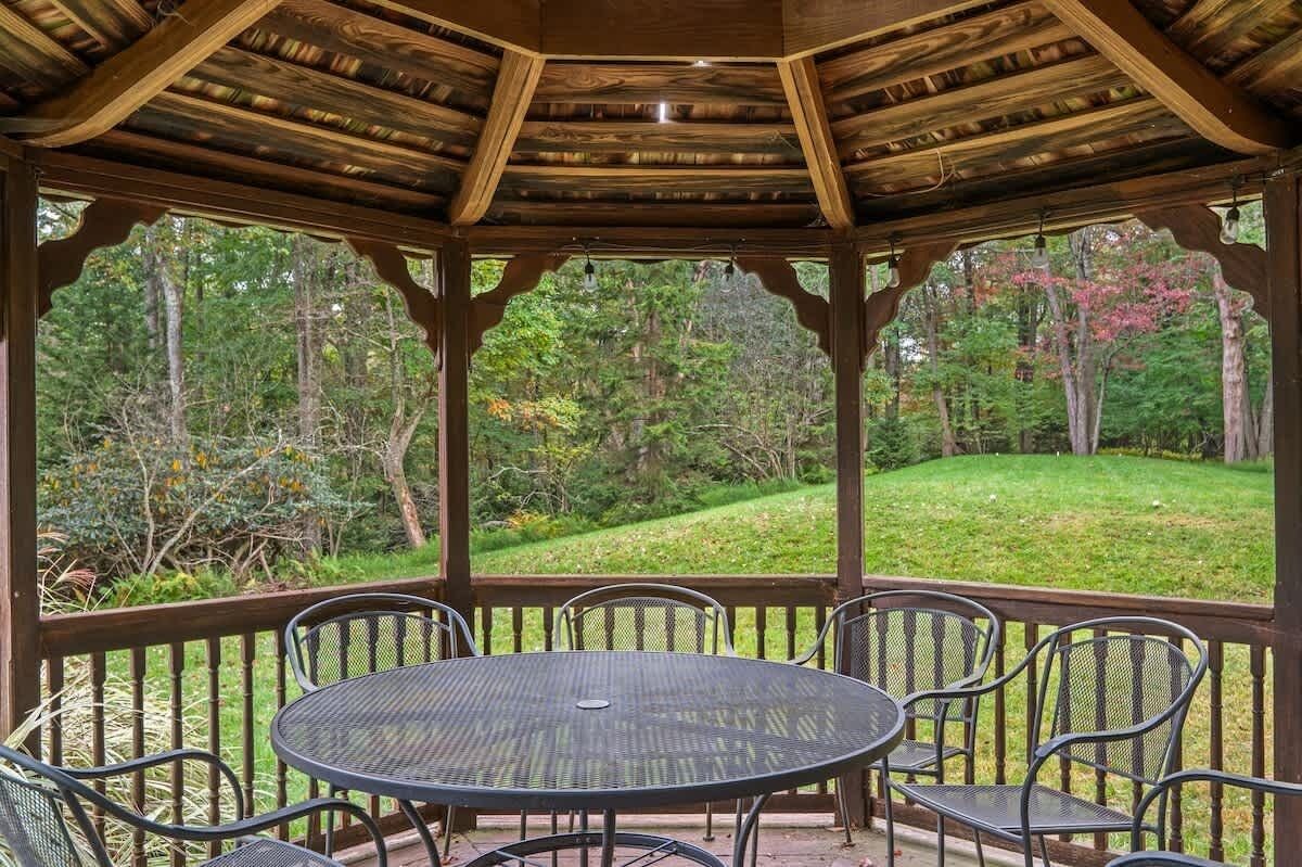 Un espace extérieur abrité, aménagé avec une table ronde et des chaises, entouré d’une terrasse en bois et offrant une vue sur un paysage boisé.