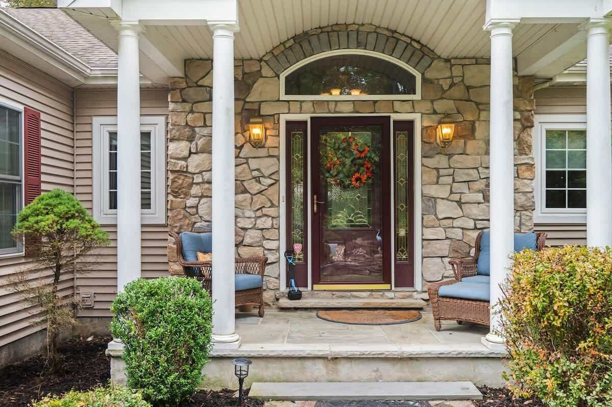 A house with a stone facade, a dark front door with a floral wreath, and two wicker chairs on the porch.