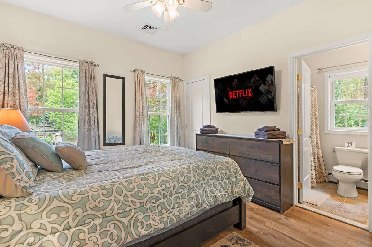 Ein Schlafzimmer mit einem Bett, einem an der Wand montierten Fernseher, einem Schminktisch und einem Fenster mit Vorhängen.