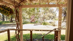 A covered porch with a wooden railing, a hanging flower basket, and a view of a park with trees and a lawn. - Secluded rental on 40 acre ranch. (Warner Springs)