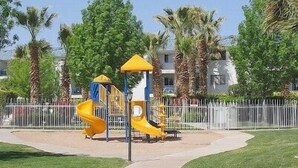 Un parque infantil con un tobogán amarillo y estructura para escalar, rodeado por una valla blanca y palmeras.