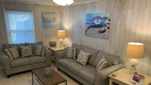 Un salon avec deux canapés gris, une table basse et une lampe. Les murs sont ornés de tableaux et il y a une fenêtre avec des stores.