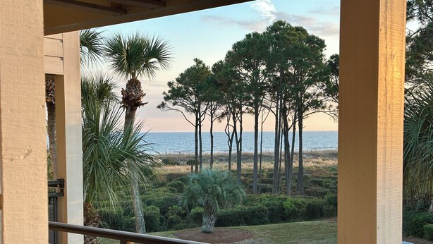 Ein Balkon mit Korbmöbeln, mit Blick auf einen Strand, hohe Bäume und einen klaren Himmel.