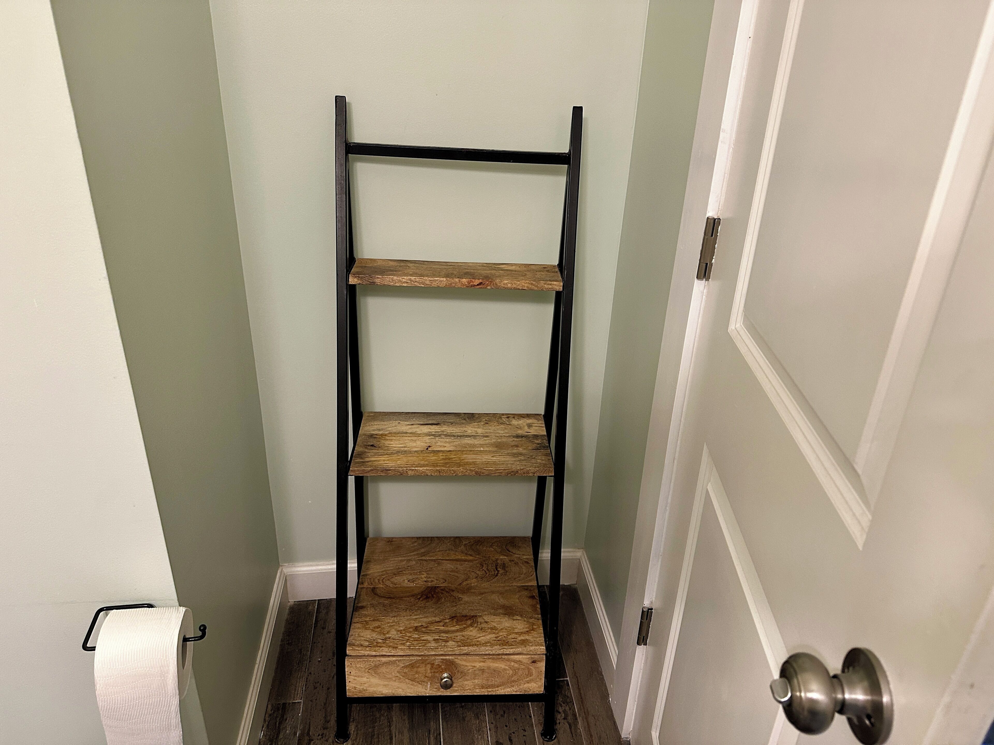 Une étagère en bois composée de trois étagères, d’un tiroir et d’une structure en métal, placée dans un couloir où l’on aperçoit une porte et un porte-rouleau de papier toilette.