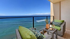Ein Balkon mit Blick auf den Ozean, Korbmöbel, ein kleiner Tisch mit einer Ananas und einer Flasche sowie ein grünes Kissen.