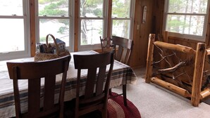 A wooden cabin interior with a dining table, chairs, and large windows offering a view of trees and a lake.