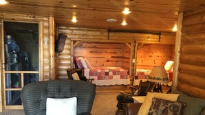 A cozy cabin interior with a wooden ceiling, a sofa with a checkered cushion, a chair with a white pillow, and a bed with a red and white checkered blanket.