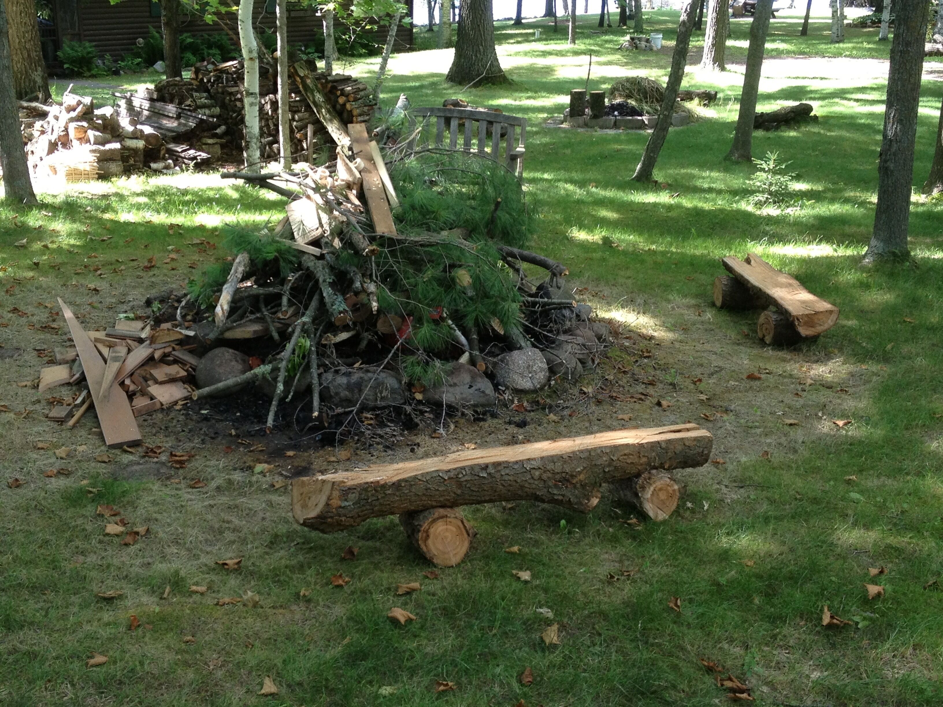 A pile of firewood and logs in a grassy area with trees and a wooden bench in the background.