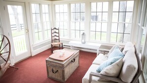 A room with a red carpet, white walls, and large windows. There is a wooden chair, a white bench, and a wicker chest of drawers.