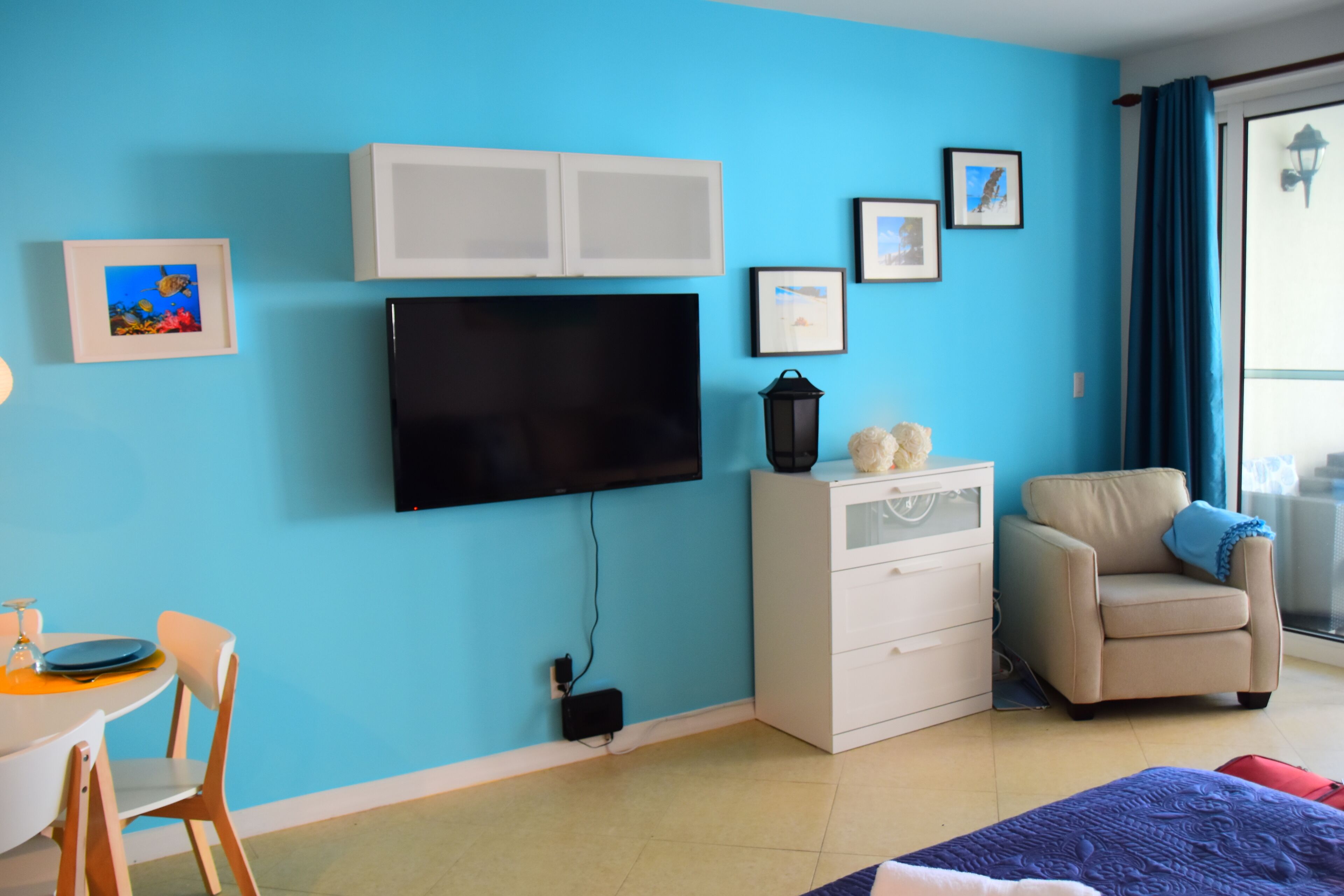 Una habitación con pared azul, un televisor de pantalla plana sobre un aparador blanco, un sillón color beige y una mesa de comedor con sillas.