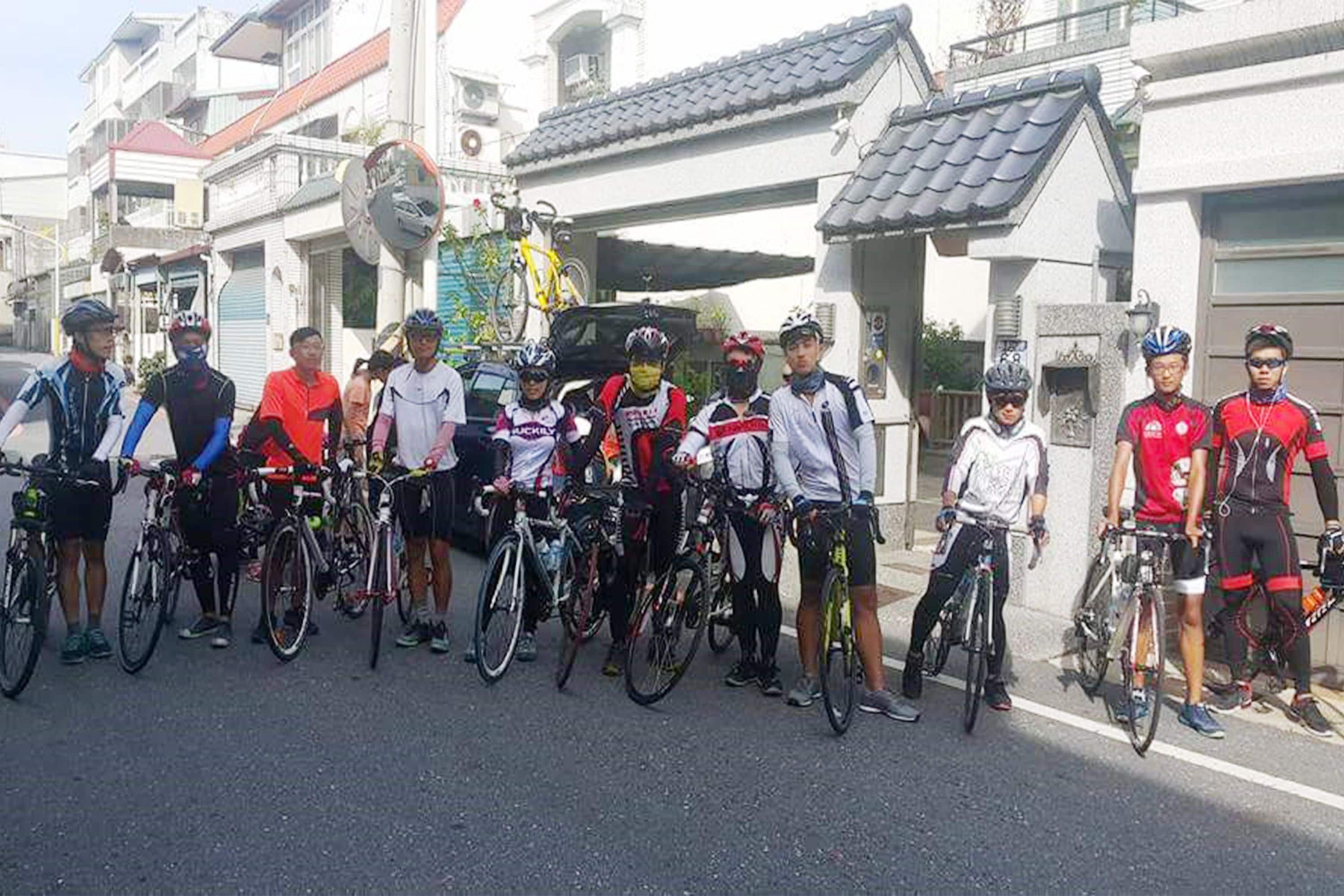 Un grupo de ciclistas de pie en fila en una calle con edificios residenciales al fondo.
