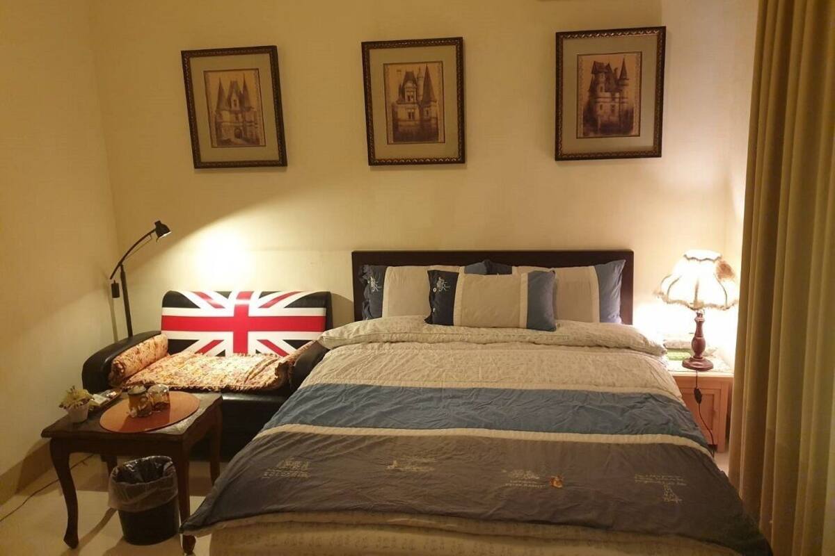 A hotel room with a bed, a chair with a Union Jack pillow, a lamp, and three framed pictures on the wall.