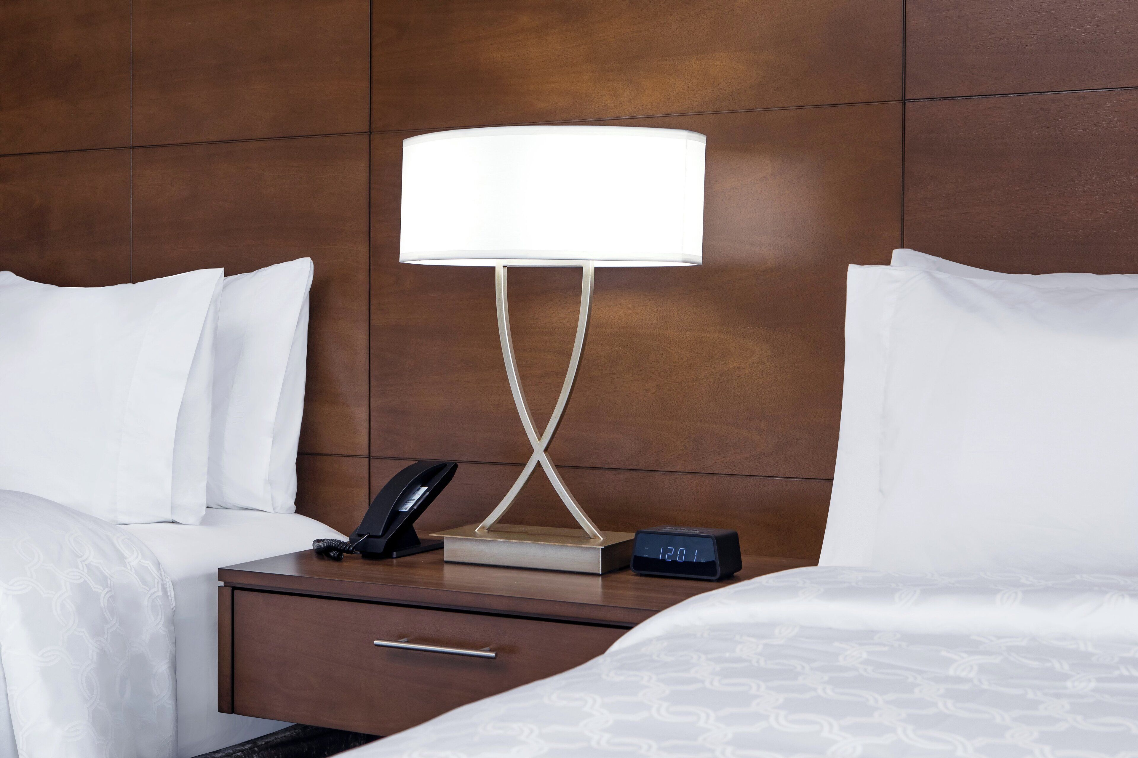 Een hotelkamer met een bed, nachtkastje, lamp, telefoon en digitale klok.