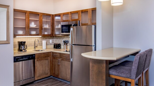 A compact kitchen with wooden cabinets, stainless steel appliances, a built-in microwave, and a small dining area with a chair. - Staybridge Suites Charlottesville Airport by IHG (Charlottesville)