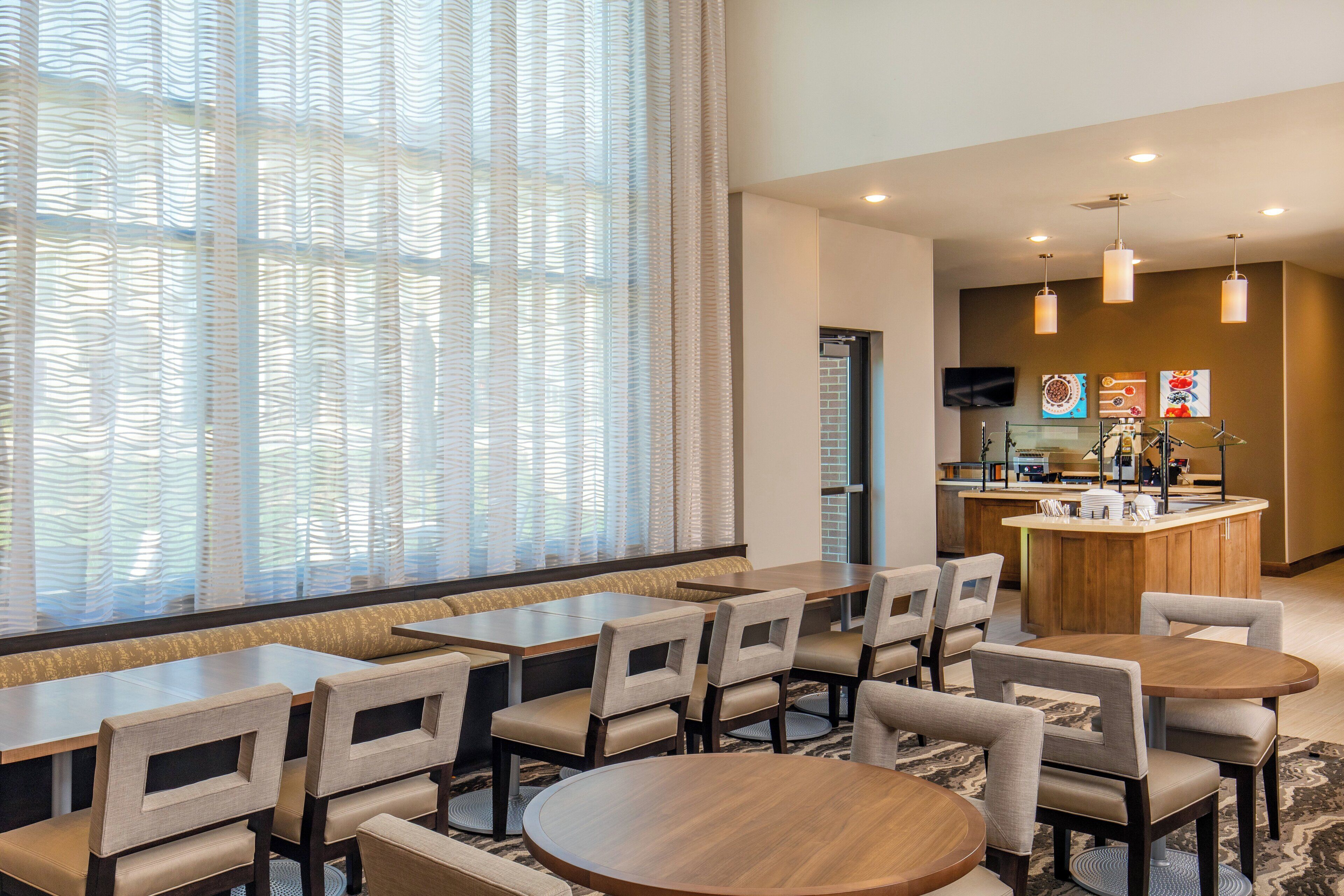 A hotel dining area with a long table, several chairs, a small round table, and a kitchen area in the background.