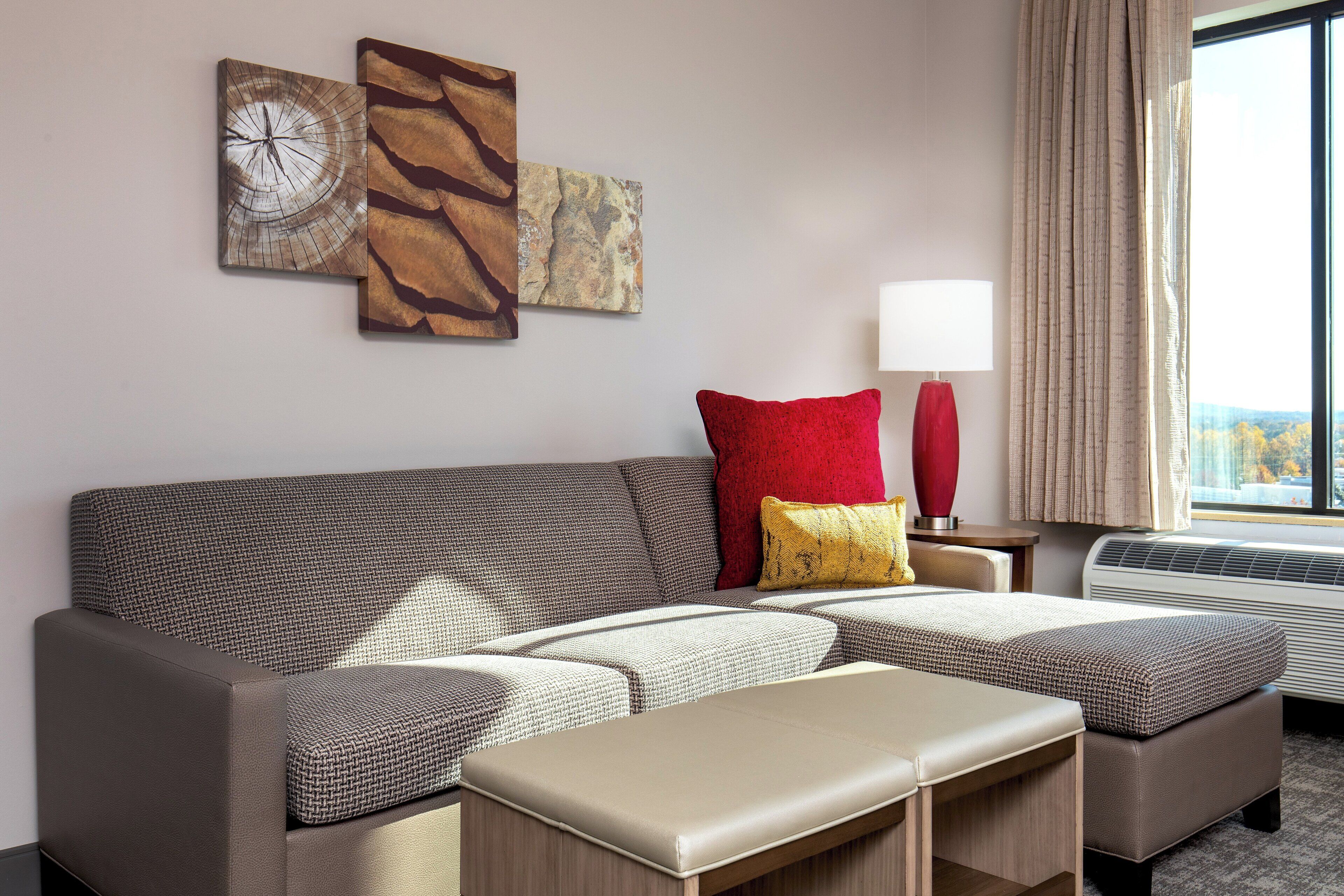 A modern hotel room with a sectional sofa, a red and yellow accent pillow, a red floor lamp, and a large window with a view.