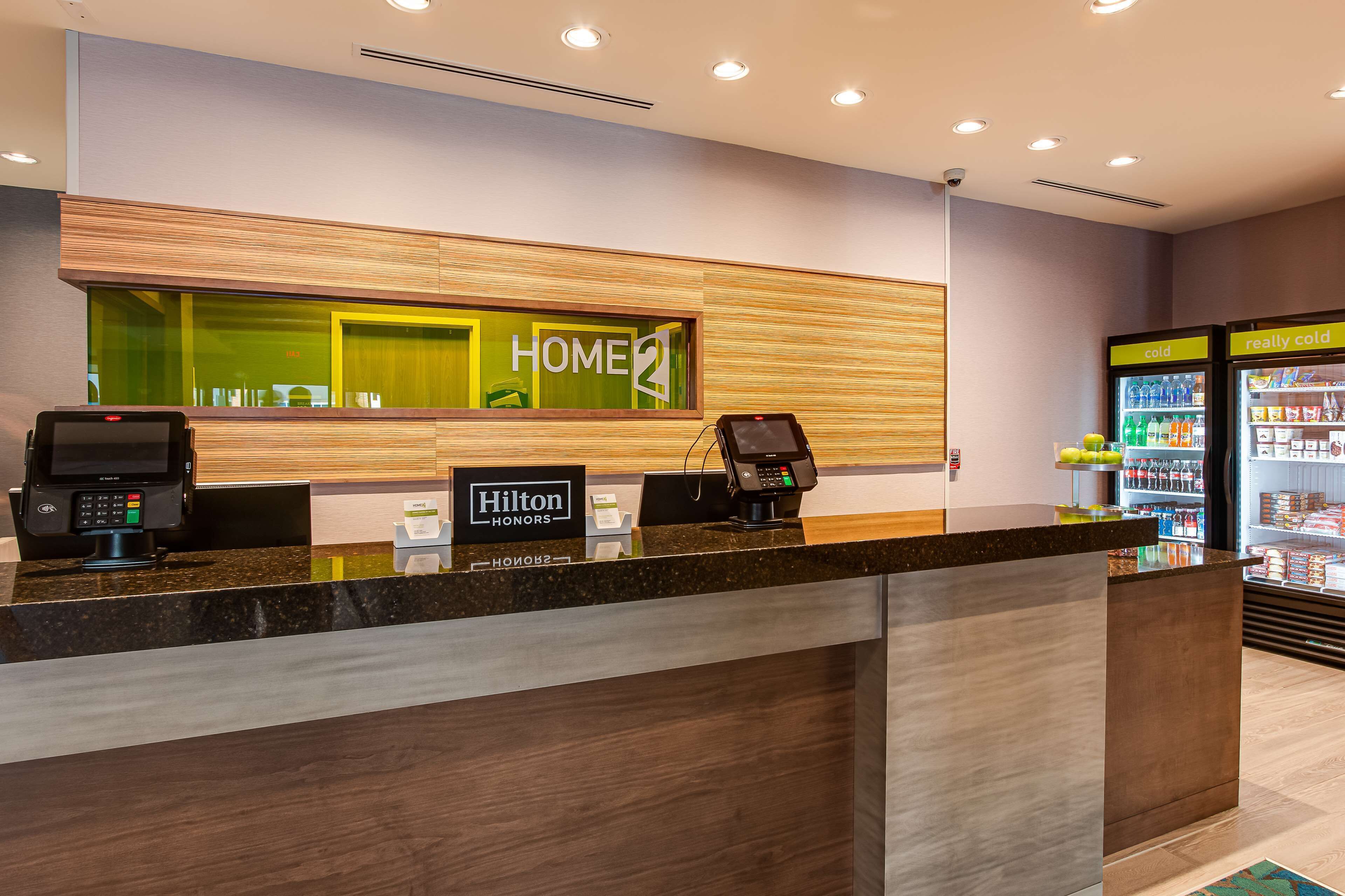 Zona de recepción de hotel con mostrador de madera, quiosco digital, minibar y nevera llena de bebidas.