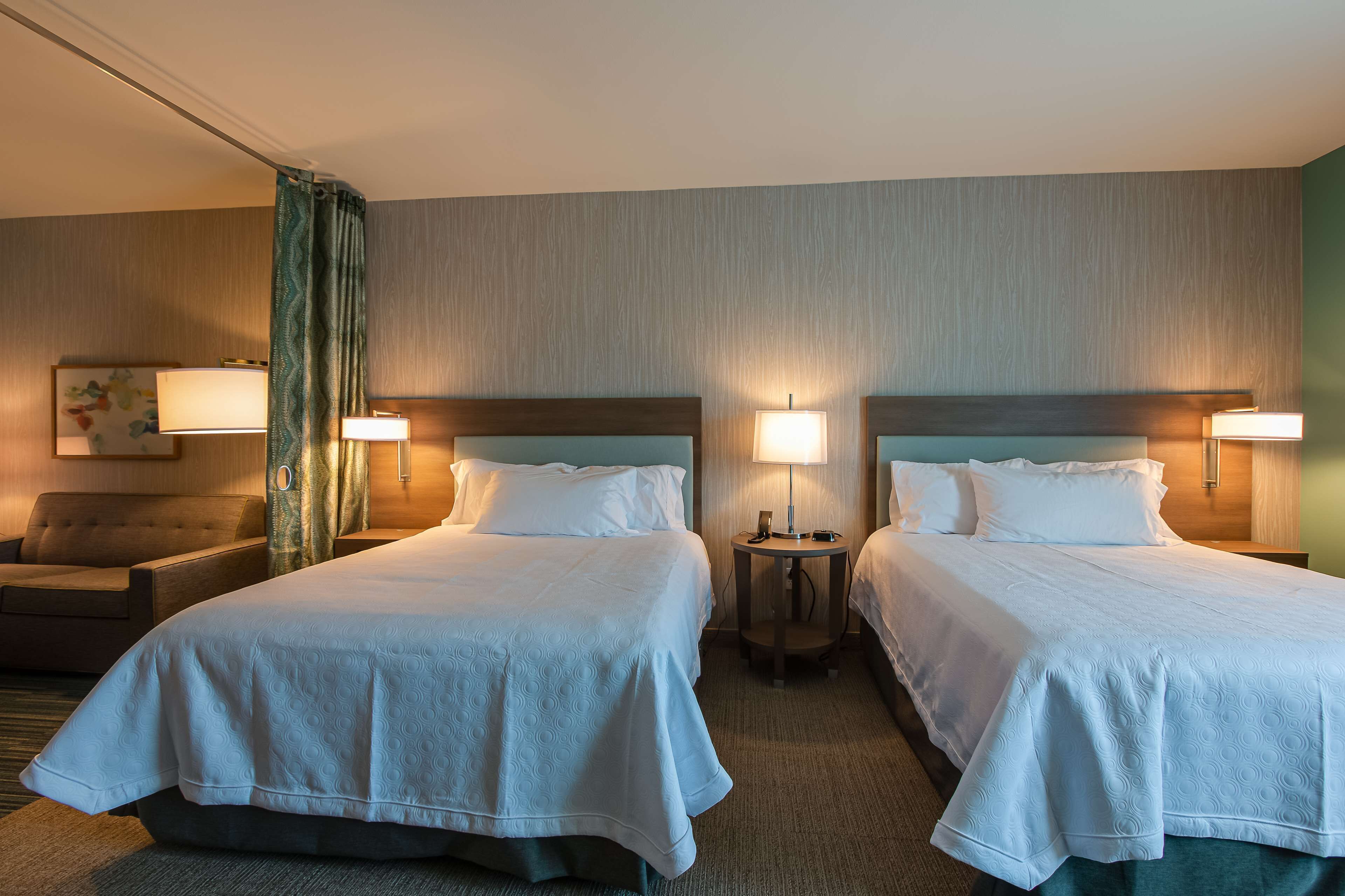 A hotel room with two beds, a small sofa, a nightstand with a lamp, and a wall-mounted picture.