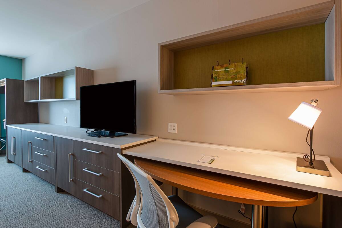 A modern hotel room with a desk, a flat-screen TV, a shelf with a book, and a desk lamp.