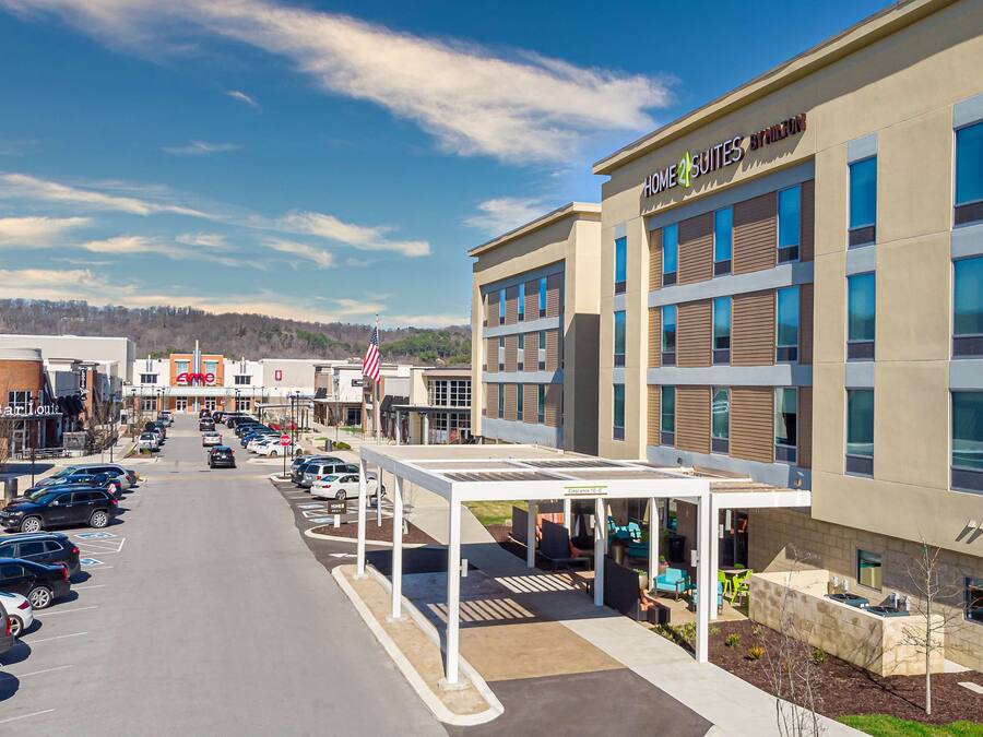 A modern hotel with a covered outdoor seating area, a parking lot, and a clear sky.