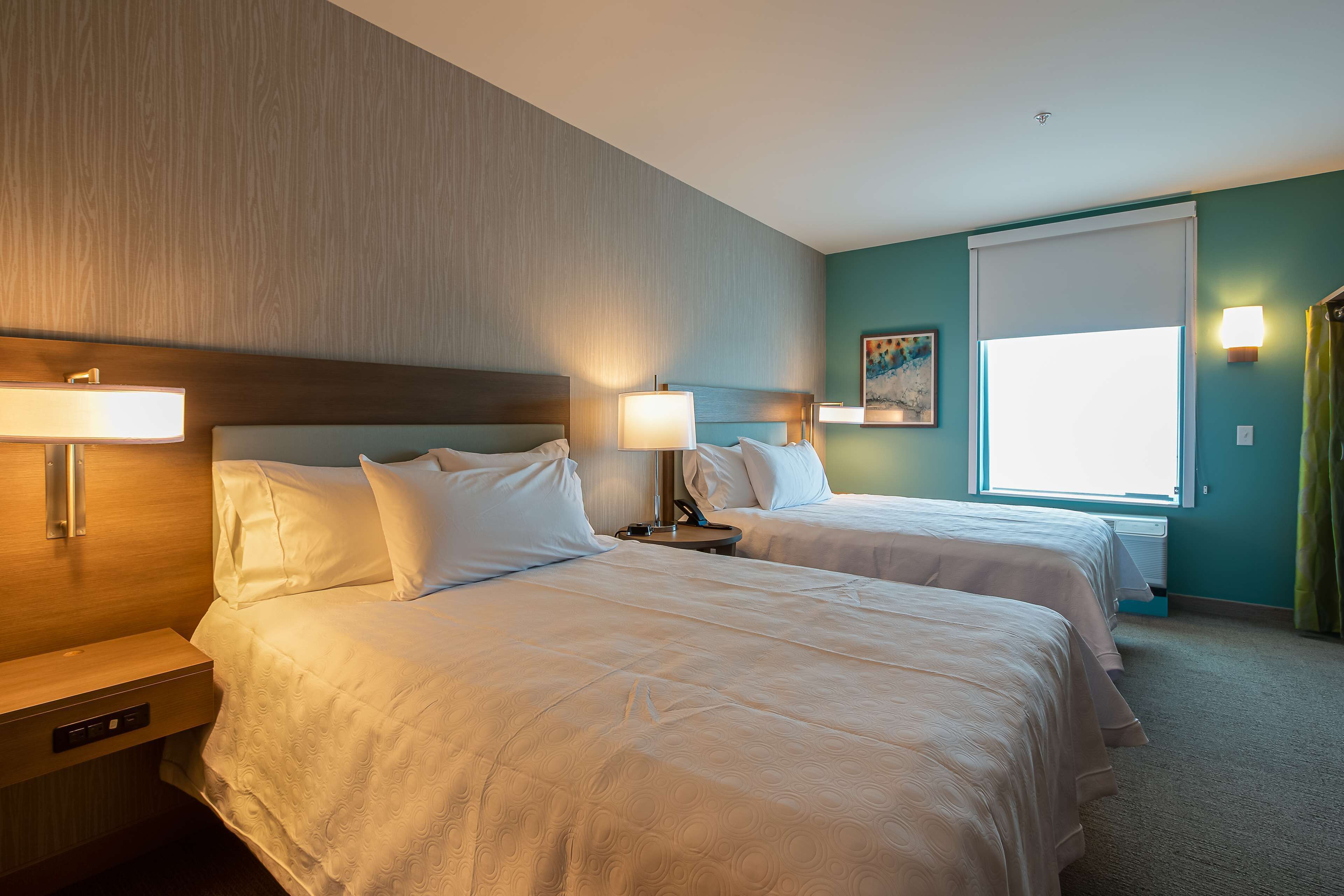 A hotel room with two beds, bedside tables, lamps, a window with curtains, and a painting on the wall.