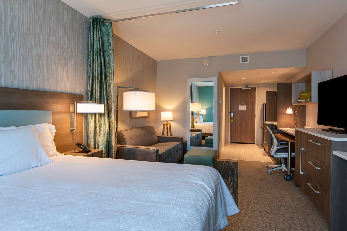 A hotel room with a large bed, a desk with a chair, a television, and a bathroom visible through an open door.