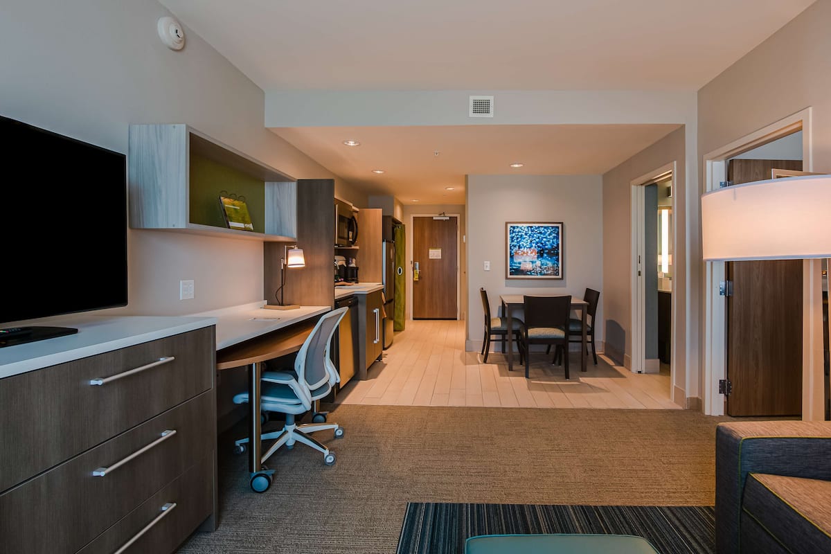 A modern hotel room with a flat-screen TV, a desk with a chair, a dining area with a table and chairs, and a large window.