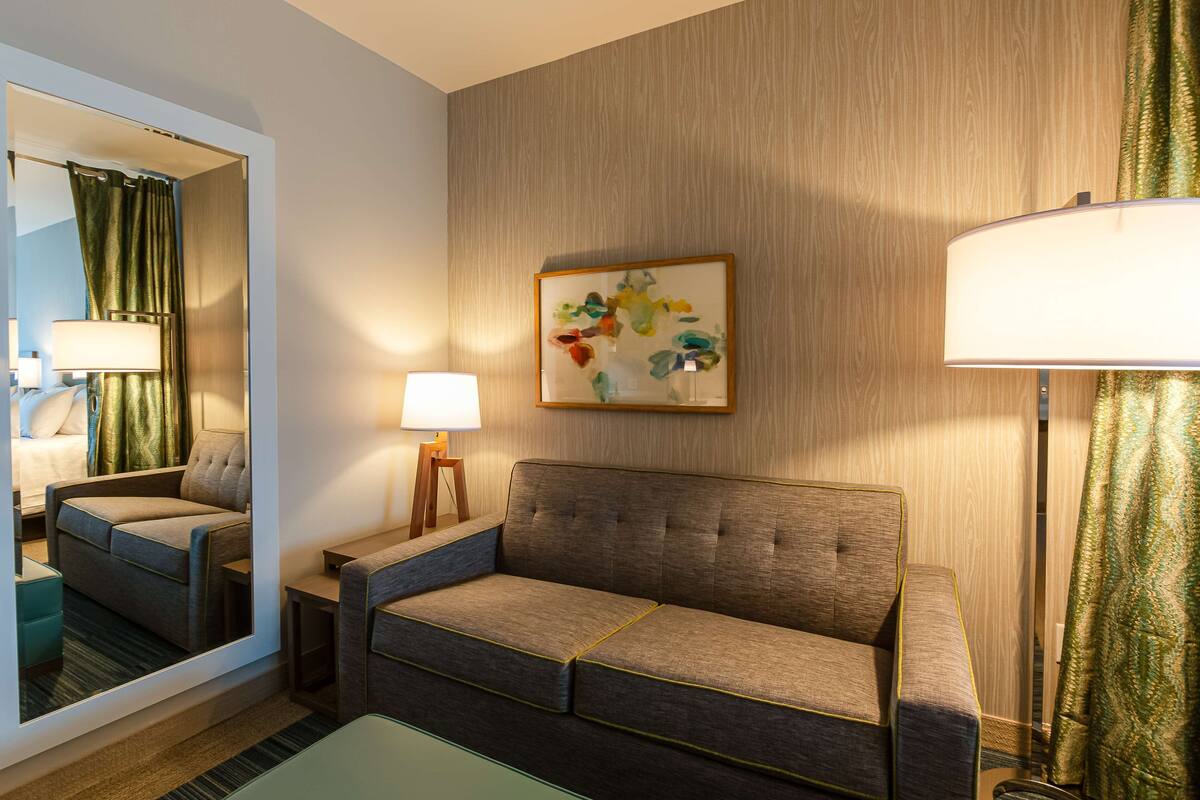 A hotel room with a sofa, a side table, a lamp, a mirror, and a painting on the wall.