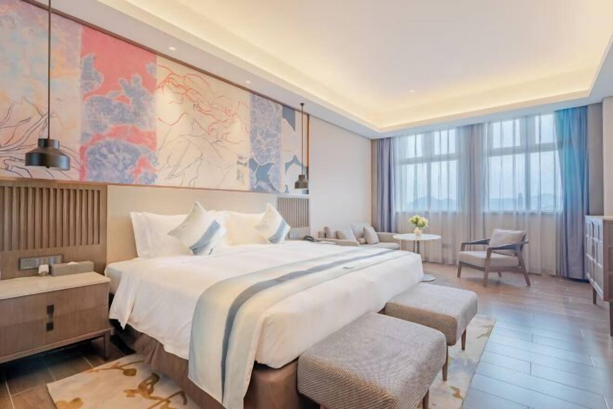 Una habitación de hotel moderna con una cama grande, una mesita de noche, una pequeña mesa redonda, una silla y un amplio ventanal con cortinas.
