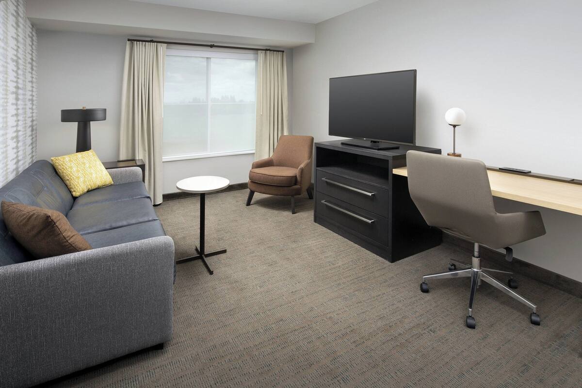 A hotel room with a grey sofa, a brown armchair, a TV, a desk, and a lamp.