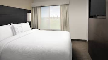 A hotel room with a large bed, a window with curtains, a flat-screen TV mounted on the wall, and a nightstand with a lamp.