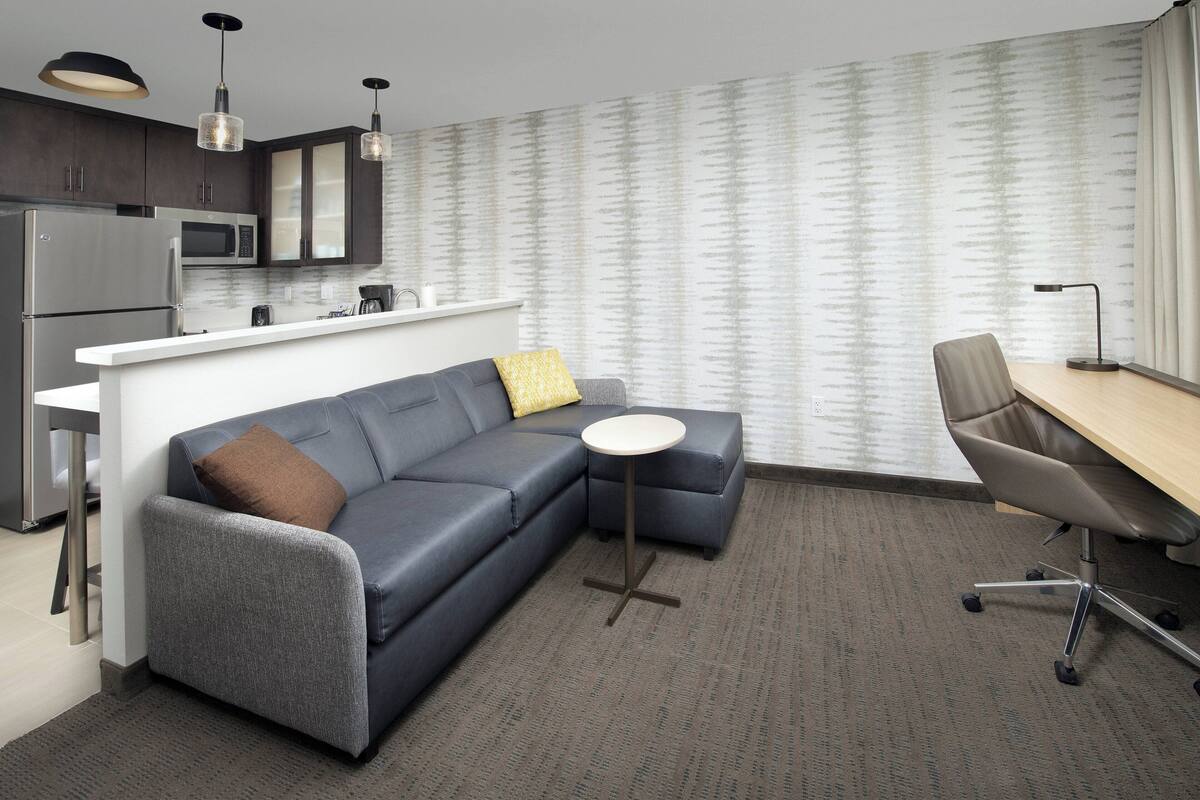 A modern kitchen with stainless steel appliances, a gray sofa with a yellow pillow, a small round table, and a wooden desk with a chair.