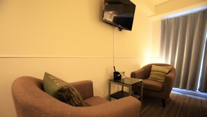 A hotel room with two sofas, a small table, a flat-screen TV mounted on the wall, and a window with curtains. - Residence Condominium KALAHAAI (Tokyo)