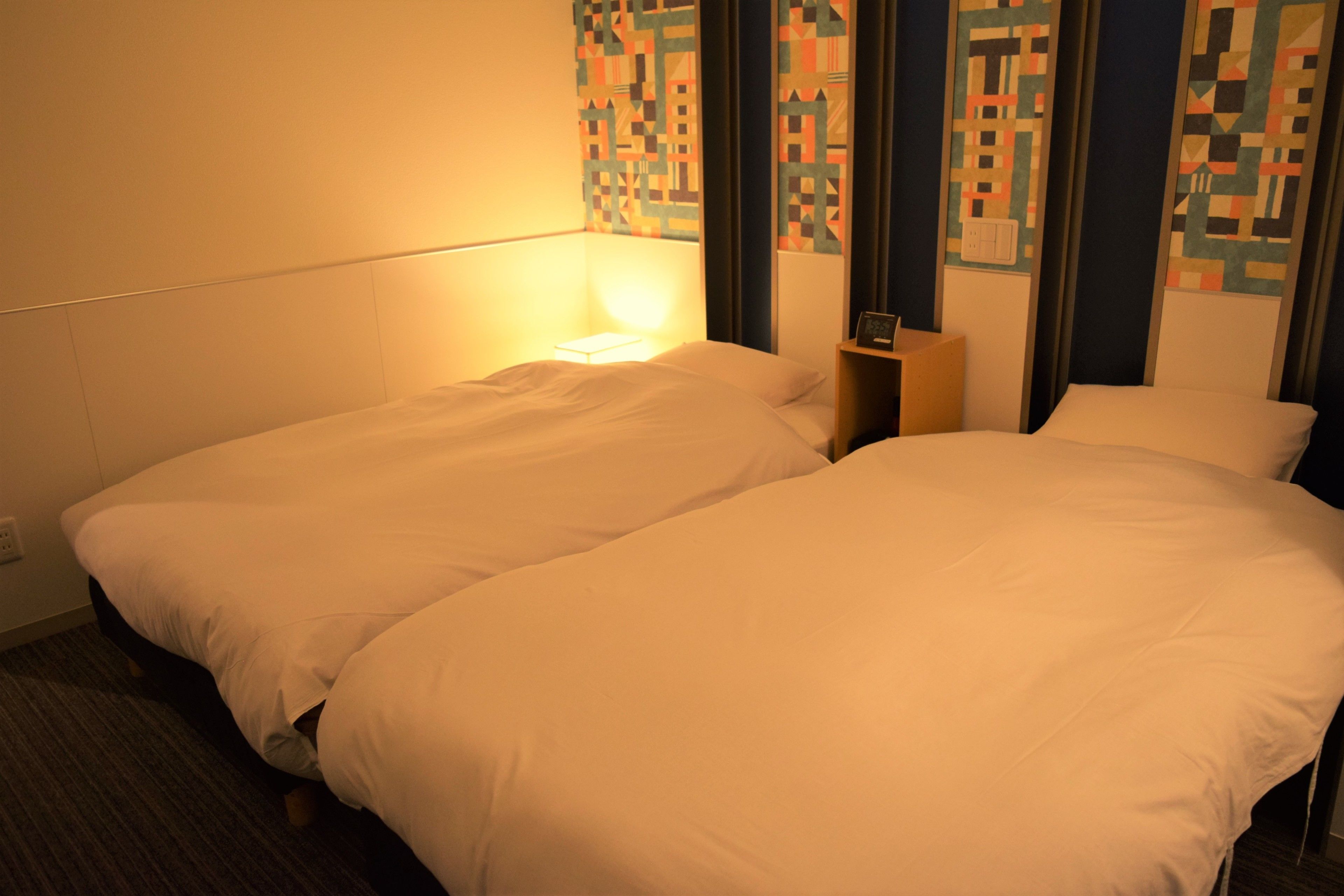 A hotel room with two single beds, a nightstand, and a wall with patterned wallpaper.