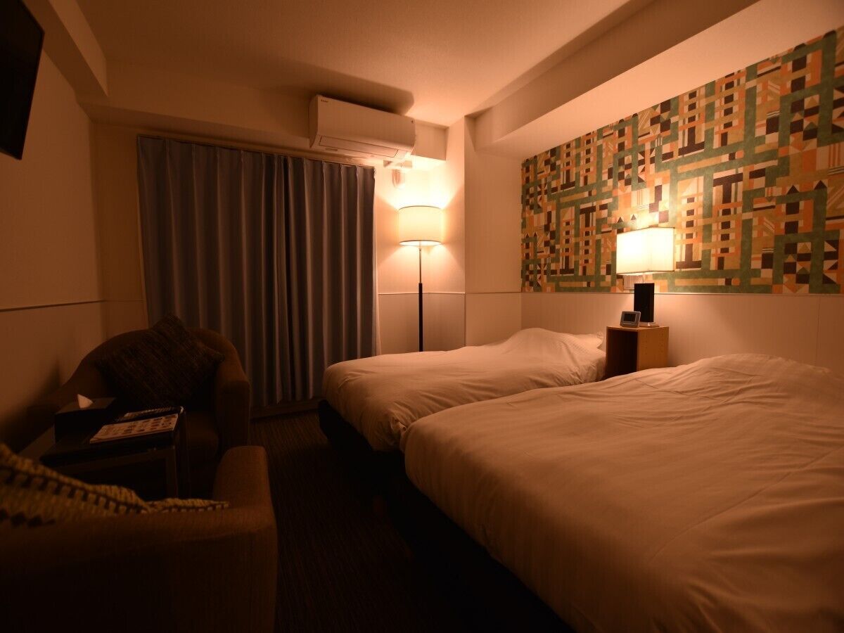 Quarto de hotel com duas camas, uma cadeira, um abajur e parede estampada.