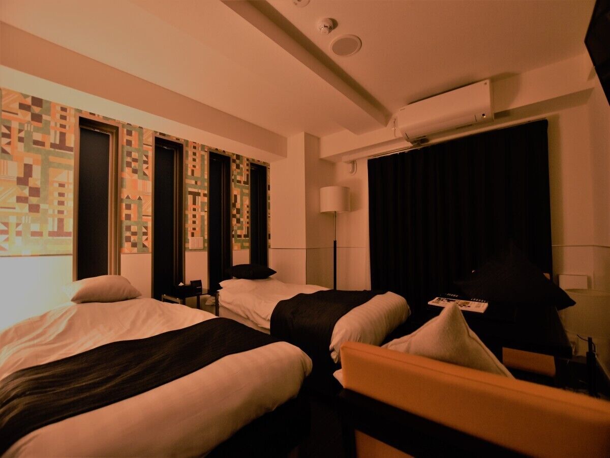 Quarto de hotel com três camas, uma escrivaninha, uma luminária e uma janela com cortinas.