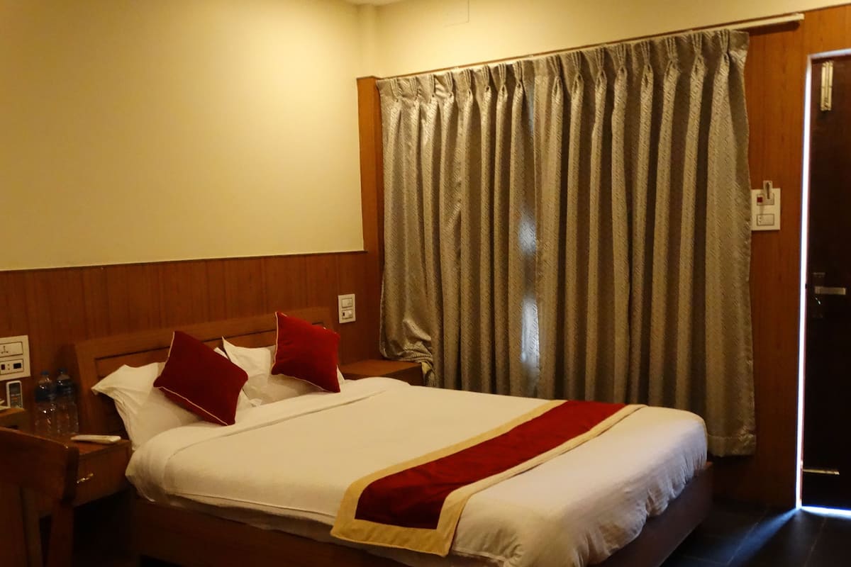 Quarto de hotel com cama de madeira, roupa de cama branca, almofadas vermelhas, manta vermelha, mesa de cabeceira com abajur e janela com cortinas.