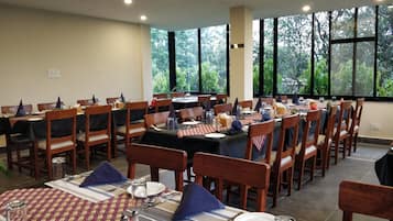 A dining area with multiple tables set for meals, wooden chairs, and large windows offering a view of greenery.