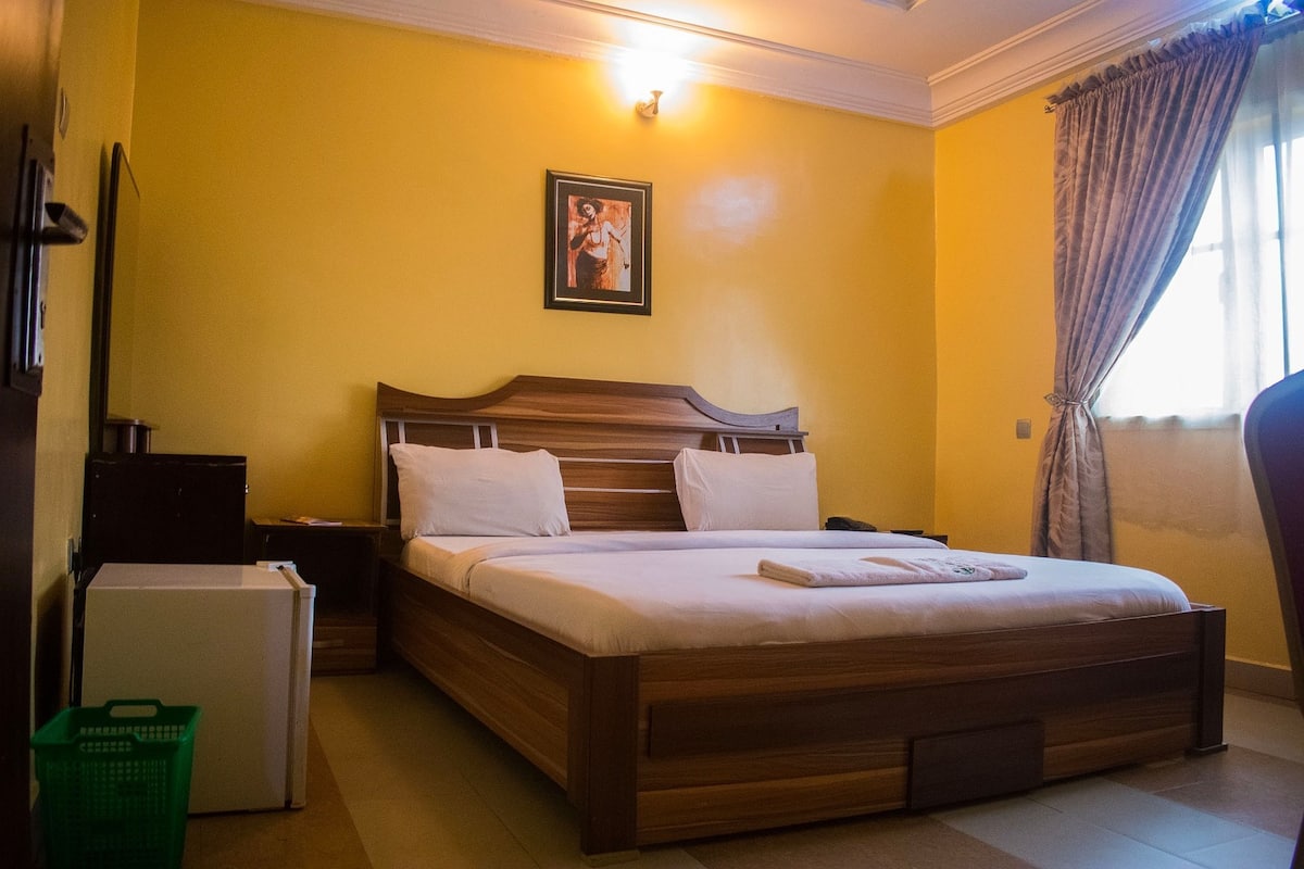 Un dormitorio con una cama de madera, almohadas blancas, una mesita de noche, un refrigerador pequeño y una cesta verde.