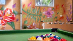 A billiards table with balls and a cue stick, set against a wall with floral artwork.