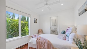 A bedroom with a bed, a ceiling fan, a window with a view of greenery, and a bathroom with a mirror and sink. - Como Palm Retreat Rye Holiday Home with Pool (Rye)
