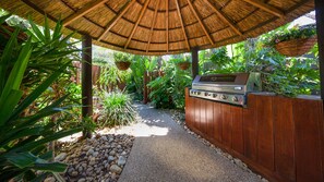 Ein Grillplatz mit Grill, umgeben von einem Holzzaun, unter einem Reetdach, mit Pflanzen und einem Kiesweg.