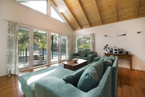 Een ruime woonkamer met een hoge plafond, grote ramen, een groene bank, een salontafel en een geluidsinstallatie.