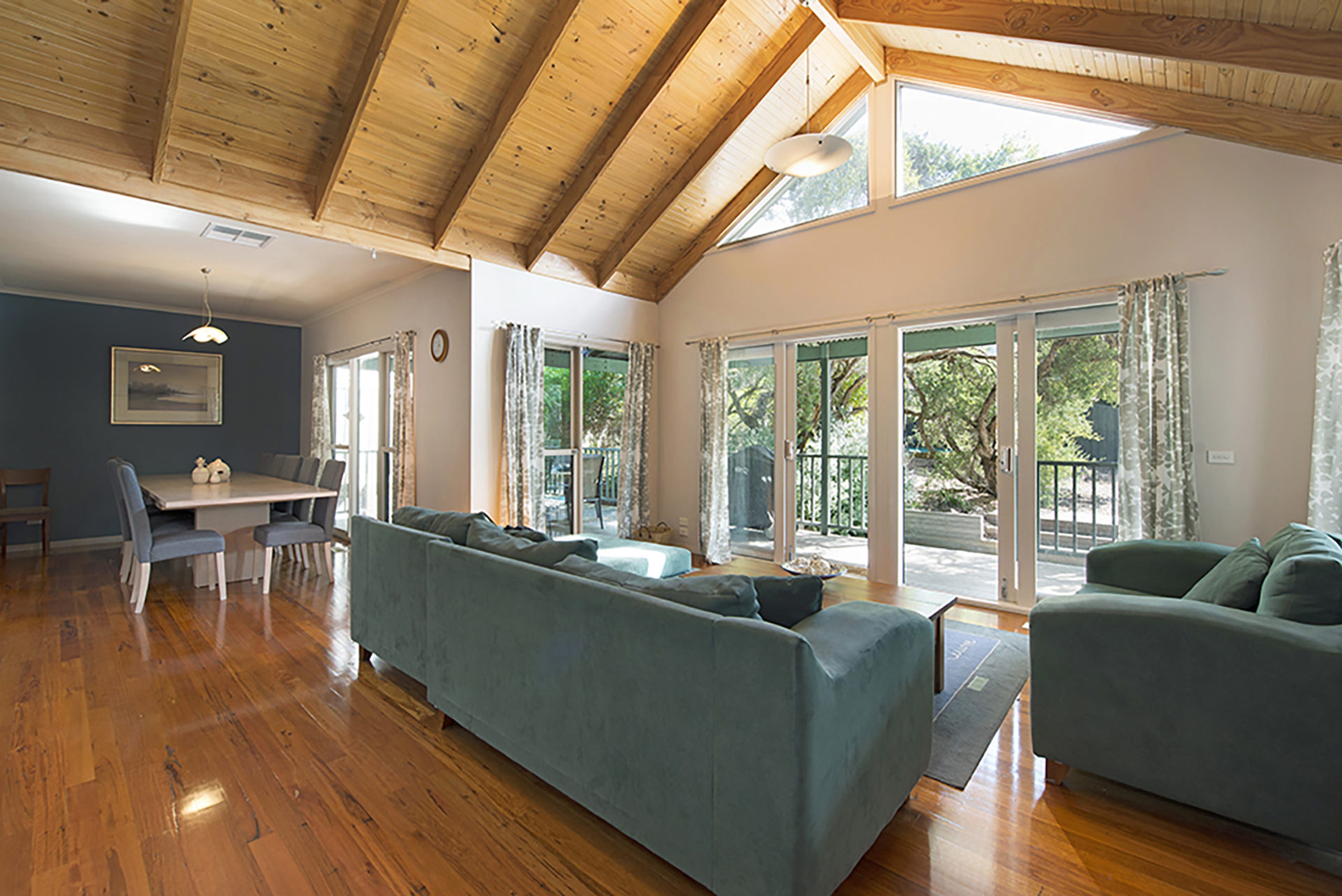 A spacious living area with a wooden ceiling, a sofa, a dining table with chairs, and large sliding glass doors leading to an outdoor area.